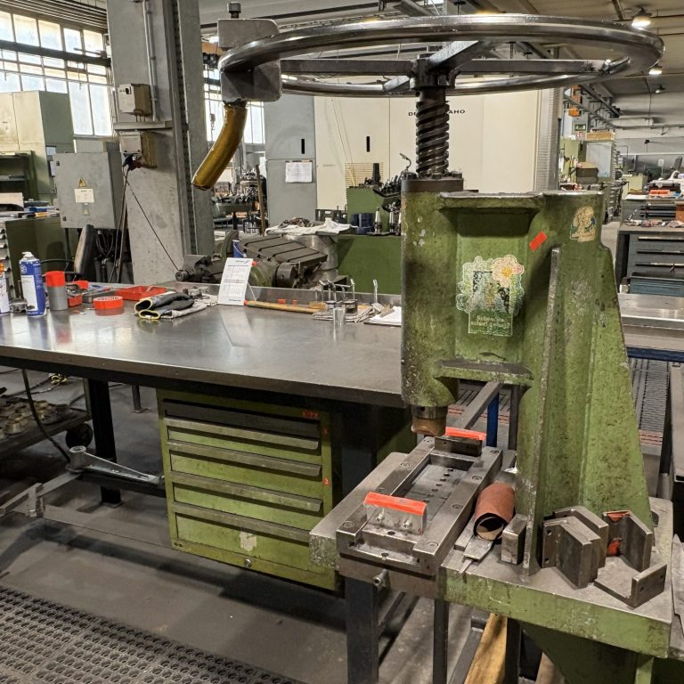 Werkbank mit Handpresse und Werkzeugen in einer Fabrikhalle.